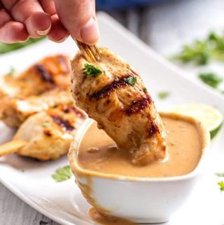 dipping a grilled chicken kabob into a small white bowl filled with peanut sauce