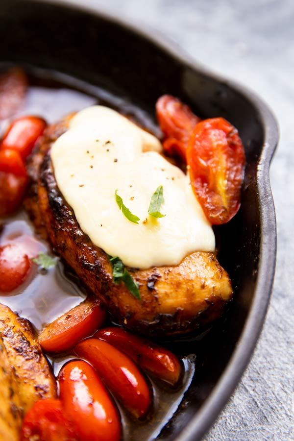 close up photo of chicken breast covered in mozzarella cheese, in a cast iron skillet with balsamic glaze and tomatoes